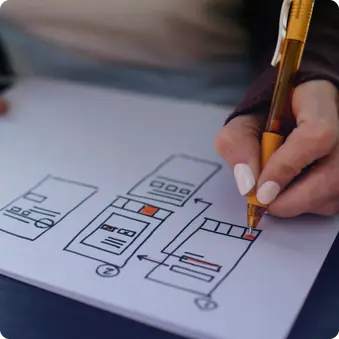 Eine Hand zeichnet mit einem orangefarbenen Stift ein schematisches Design auf einem Blatt Papier. Das Design zeigt mehrere Rechtecke und Pfeile, die verschiedene Abschnitte und Navigation darstellen.